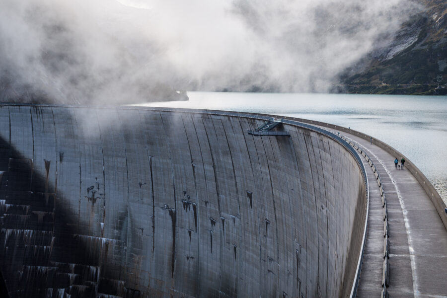 Massive dam wall in the middle of a mountain range, surrounded by clouds.