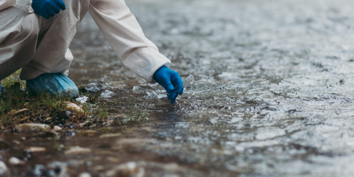 Wissenschaftler im Schutzanzug entnimmt Wasserproben aus einem Fluss zur ökologischen Analyse.