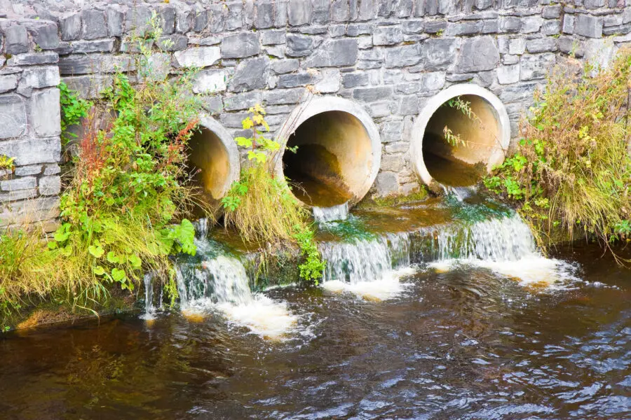 Giftiges, verschmutztes Wasser fließt aus drei Betonabflussrohren in einen Fluss, umgeben von Vegetation.