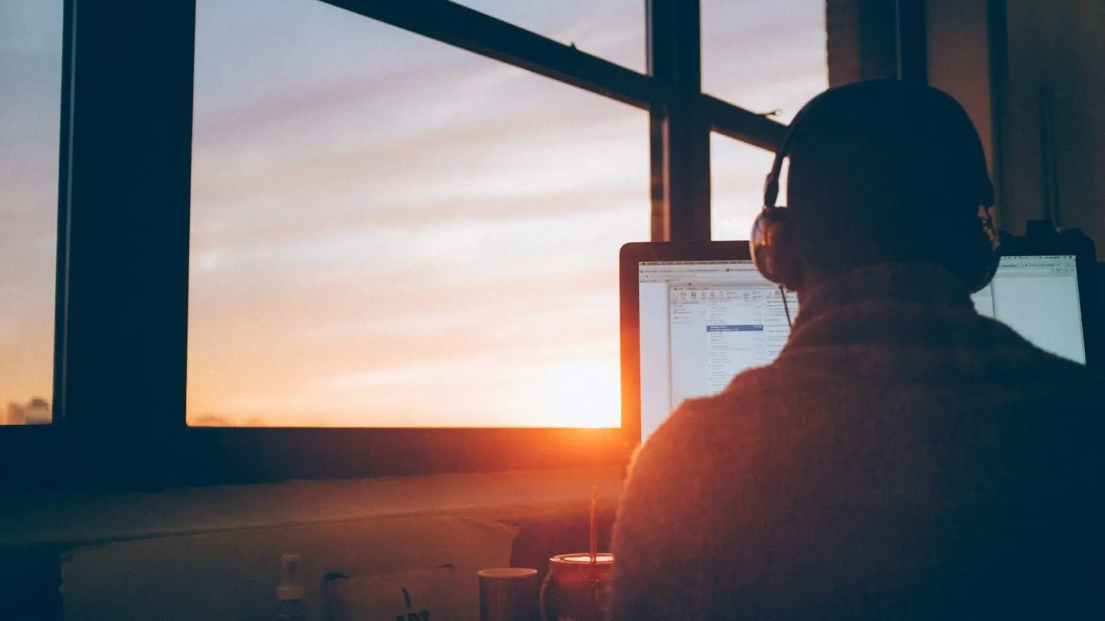 Man with headphones sitting in front of a computer at his workstation next to a large window as the sun sets.