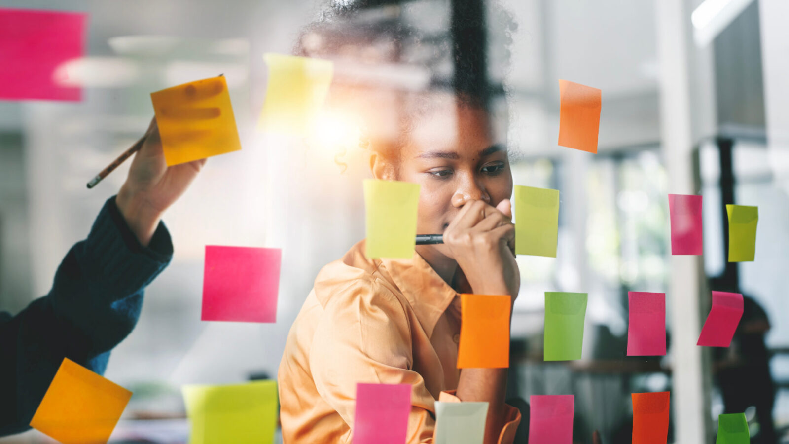 Young businesswoman writes on a coloured sticky note on a glass wall, while another person also writes down ideas on a note.