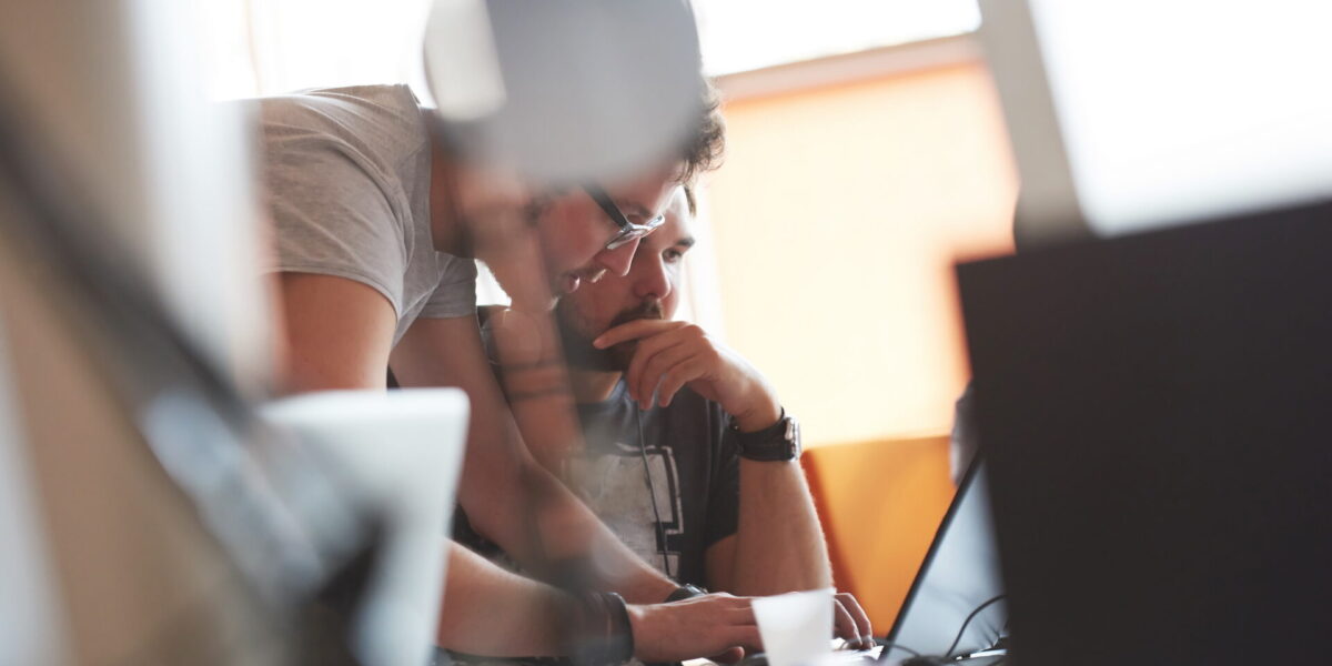 In a modern office, a man explains something to his colleague on the computer, both of them focussed on the screen.