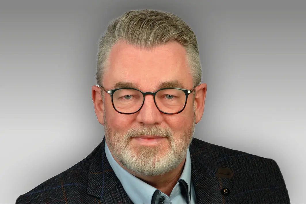 Ein Mann mittleren Alters mit grauem Haar, Bart und Brille trägt eine dunkle Jacke und ein hellblaues Hemd und blickt vor einem grauen Hintergrund mit Farbverlauf direkt in die Kamera.