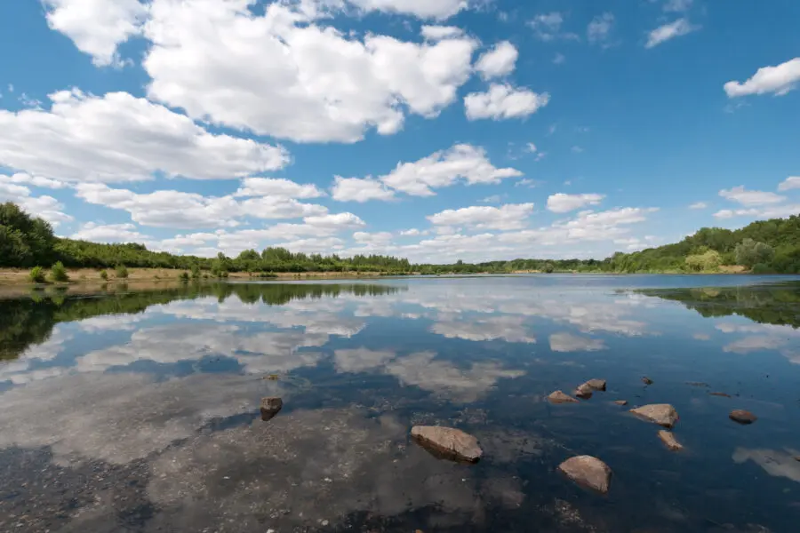 Ein ruhiger See mit klarem Wasser, in dem sich ein teilweise bewölkter blauer Himmel spiegelt, umgeben von grünen Bäumen und grasbewachsenen Ufern, mit Felsen im flachen Vordergrund.