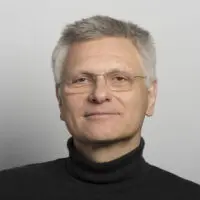 Ein Mann mittleren Alters mit kurzen grauen Haaren und Brille, der einen schwarzen Rollkragenpullover trägt und sanft vor einem schlichten hellgrauen Hintergrund lächelt.