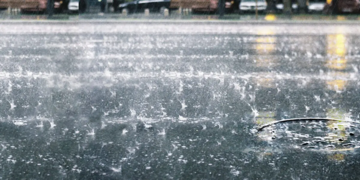 Heftiger Regen prasselt auf eine Straße, während Wasser durch einen Kanaldeckel abfließt.