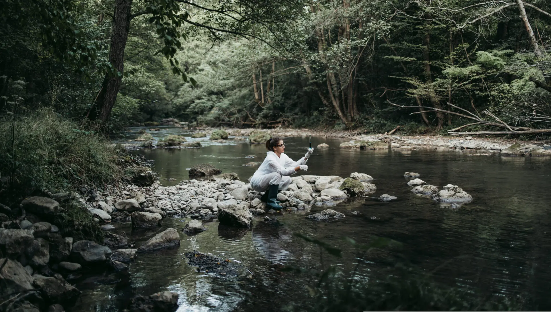 Wissenschaftlerin in Schutzkleidung entnimmt eine Wasserprobe in einem natürlichen Flusslauf, um die Wasserqualität zu analysieren.