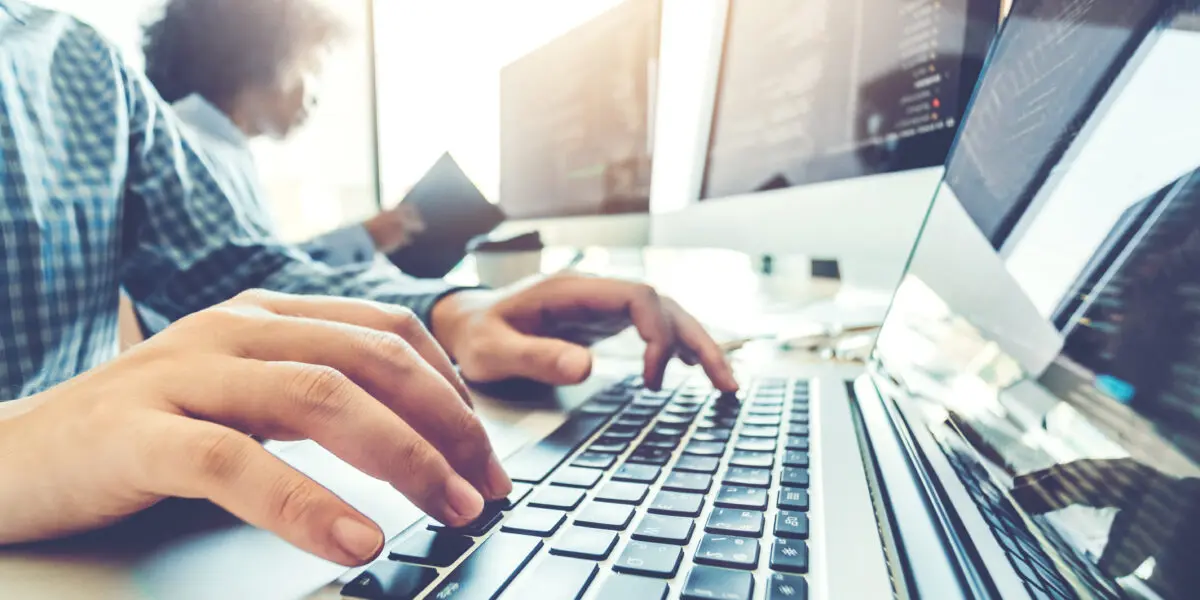 Close-up of hands on a keyboard, several monitors, in the background a woman concentrates on her office work.