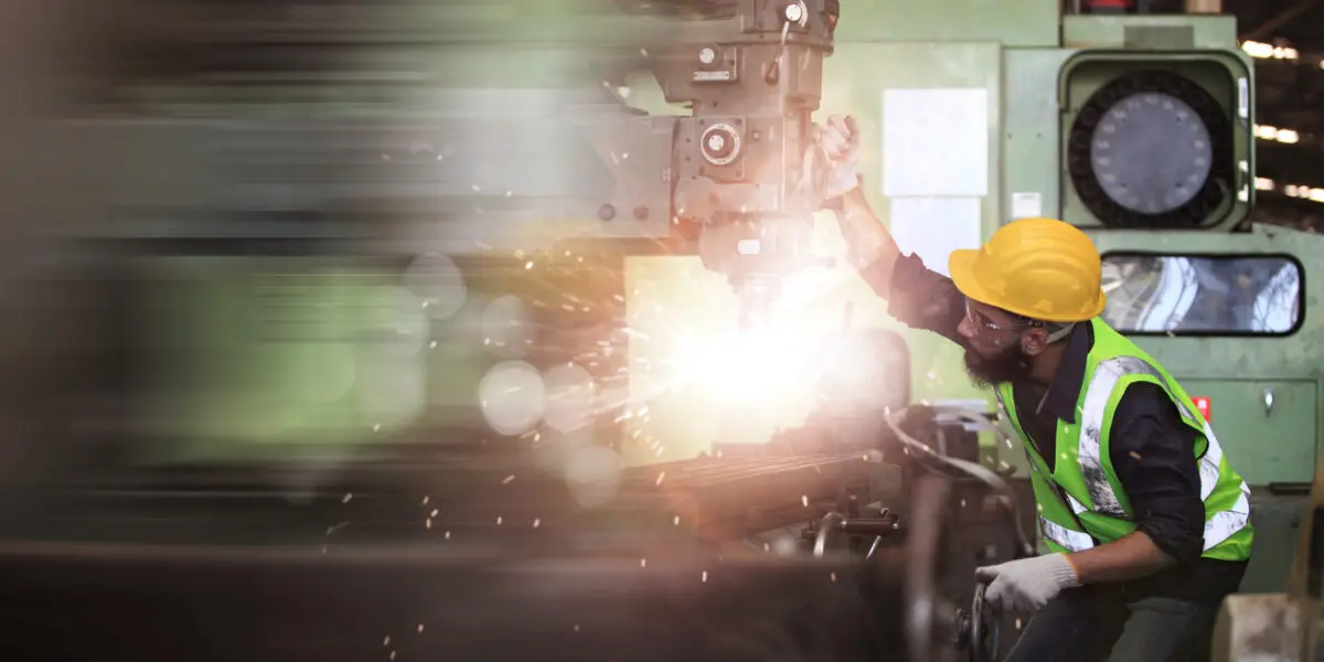 A factory worker wearing a yellow hard hat and high-visibility waistcoat operates a heavy industrial drill while sparks fly.