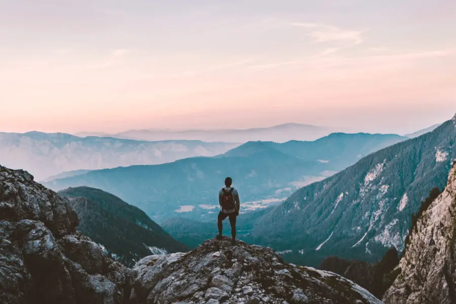 Wanderer mit Rucksack steht auf einem Felsen und blickt über ein weites Bergpanorama bei Sonnenaufgang.
