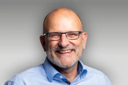 Ein lächelnder Mann mittleren Alters mit Glatze, Brille und gestutztem grauen Bart, der ein hellblaues Hemd mit Kragen trägt, posiert vor einem hellgrauen Hintergrund mit Farbverlauf.
