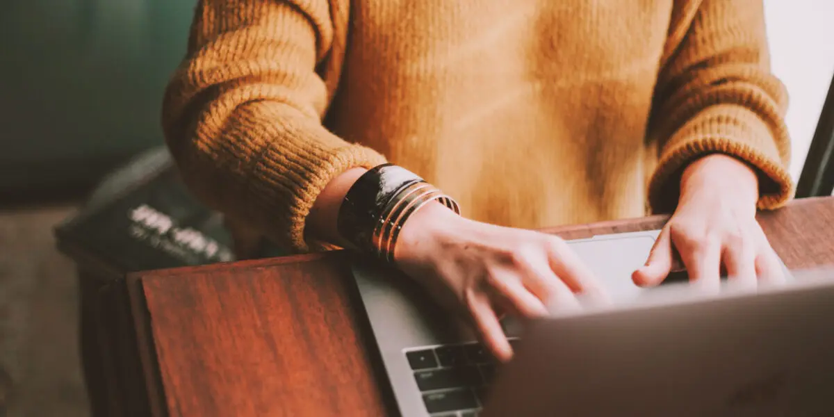 Person in a mustard yellow jumper types on a laptop at a wooden table while natural light shines through the window.