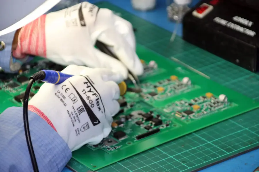 A person wearing white safety gloves is soldering electronic components onto a green circuit board using a soldering iron on a workbench with a green cutting mat.