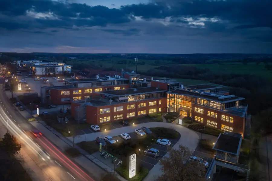 Beleuchtetes Bürogebäude bei Dämmerung mit moderner Architektur und Parkplatz im Vordergrund.