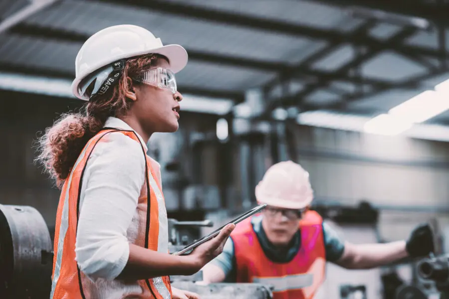 Industrieingenieurin mit weißem Helm und Schutzbrille in einer Produktionshalle, während ein Mitarbeiter eine Maschine bedient.