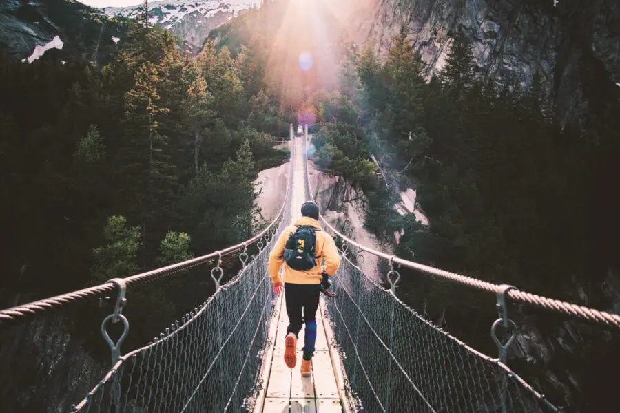 Mann mit Rucksack und Outdoorkleidung überquert eine Hängebrücke inmitten einer Schlucht, umgeben von Bergen und Bäumen.