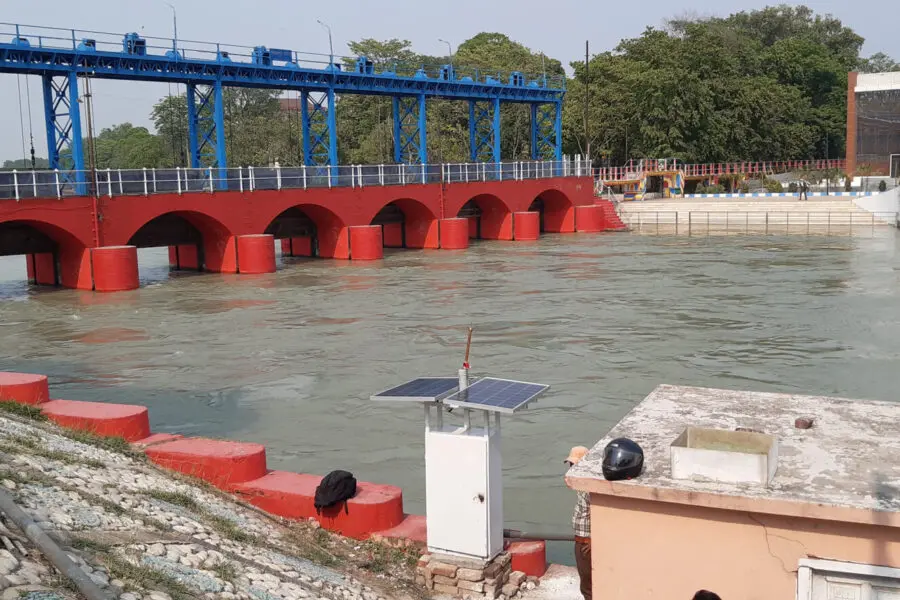 A blue and red water control structure spans a flowing river near a building, surrounded by trees and a clear sky. A small solar panel and rooftop are visible in the foreground.