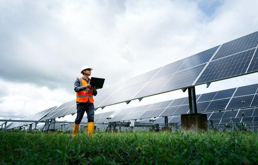 A person in a safety vest and helmet stands on grass near a large array of solar panels, holding a laptop and looking at the panels, with a cloudy sky in the background.
