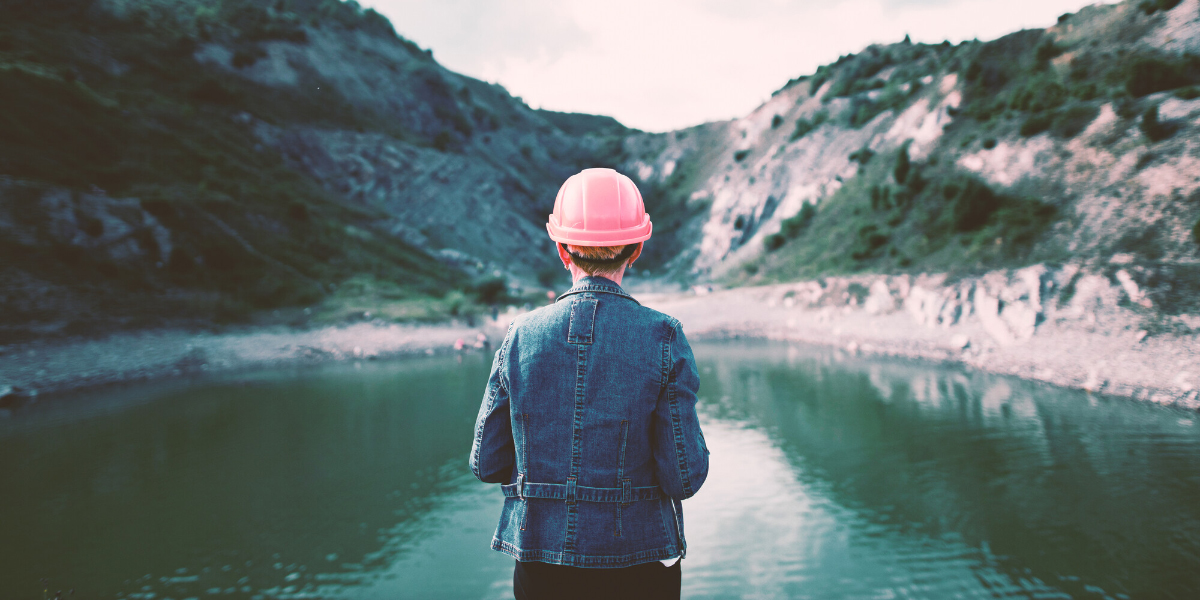 A person wearing a pink hard hat and a denim jacket stands at the edge of a calm lake and looks out over rugged green hills.