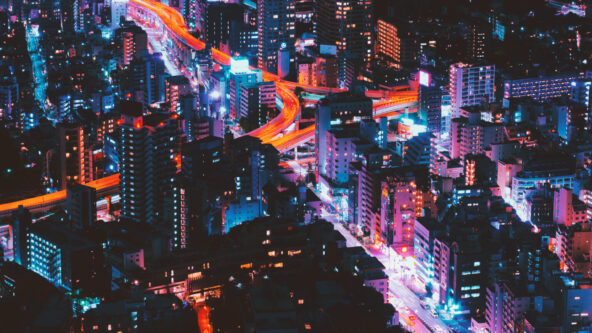 Eine lebendige Stadtlandschaft bei Nacht mit hohen, neonbeleuchteten Gebäuden und leuchtenden Fenstern. Helle, gewundene Straßen erzeugen leuchtende rote und blaue Streifen, die das dicht besiedelte Stadtgebiet durchziehen.