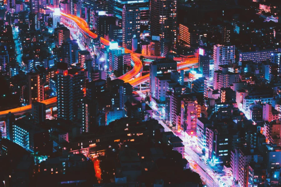 A lively cityscape at night with tall, neon-lit buildings and glowing windows. Bright, winding streets create glowing red and blue stripes.