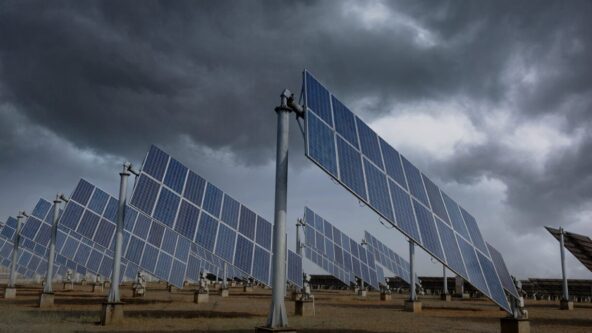 A person in a safety vest and helmet stands on grass near a large array of solar panels, holding a laptop and looking at the panels, with a cloudy sky in the background