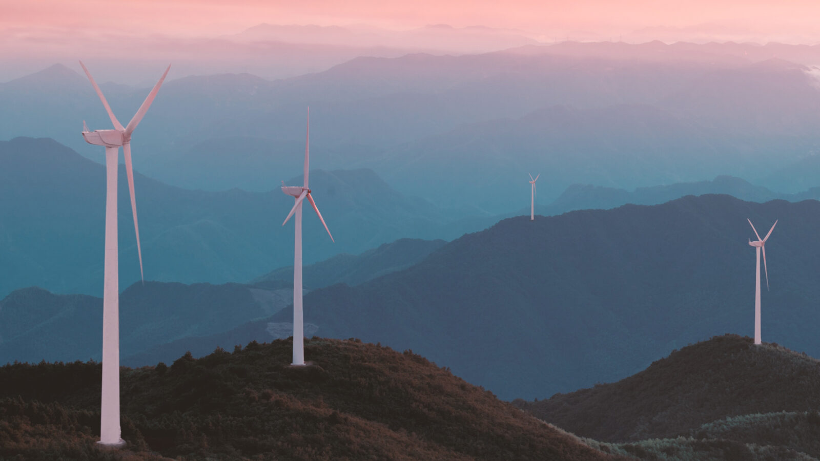 Vier Windturbinen stehen auf grünen Bergkämmen unter einem rosafarbenen und violetten Himmel bei Sonnenuntergang.