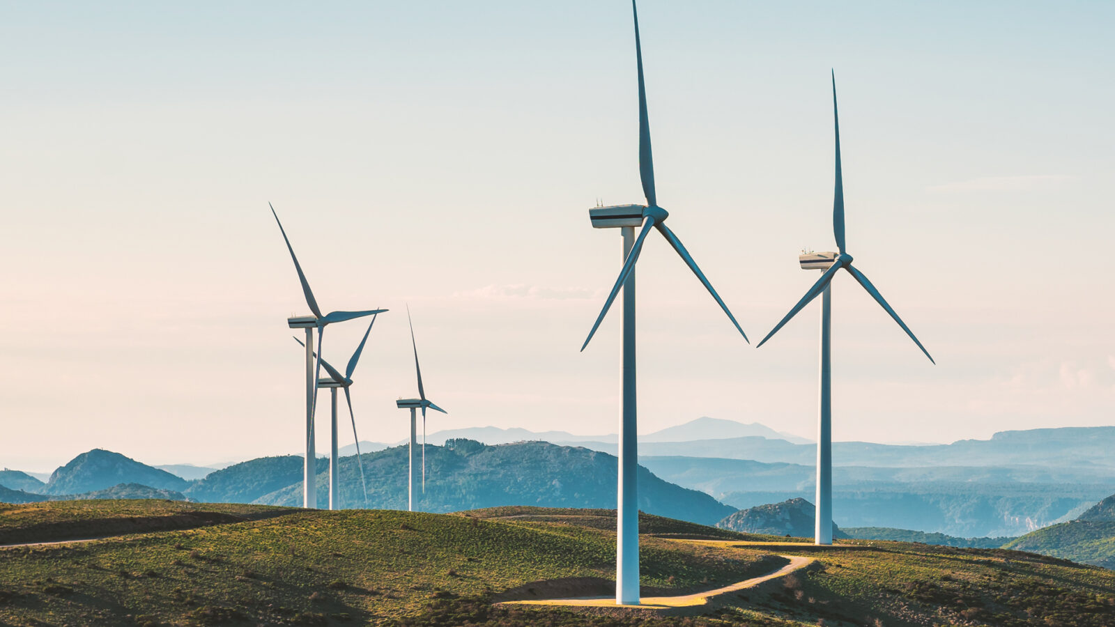 Große Windturbinen stehen auf einem grasbewachsenen Hügel und erzeugen erneuerbare Energie. Im Hintergrund sind sanfte Hügel und ferne Berge.