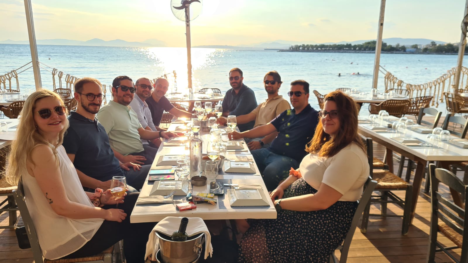 A group of ten people sit around a table at an outdoor seaside restaurant at sunset, smiling and enjoying drinks together. The sea and sun are visible in the background.