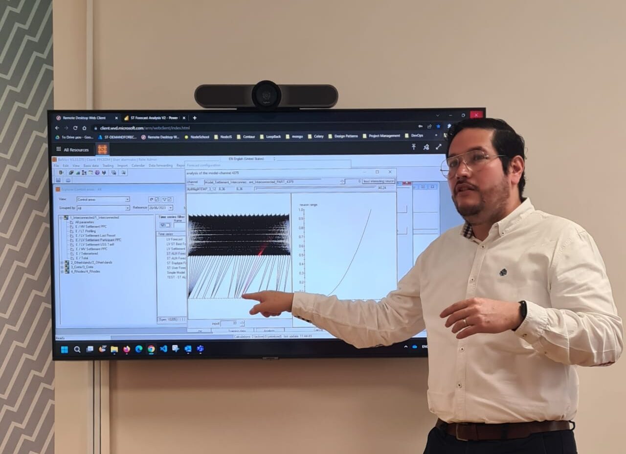 A man in a white shirt is standing and pointing at a large screen displaying graphs and data charts in a classroom or office setting. The screen shows a black-and-white plot and a curved line graph.