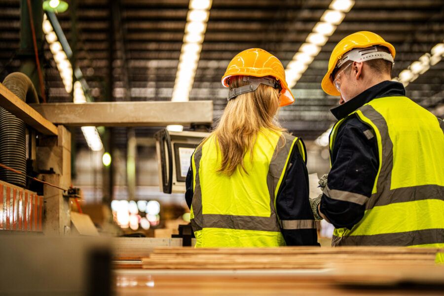 Zwei Arbeiter mit gelben Sicherheitswesten und orangefarbenen Schutzhelmen stehen in einer Fabrik vor Maschinen und Computerbildschirmen, im Vordergrund sind Stapel von Holzbrettern zu sehen.