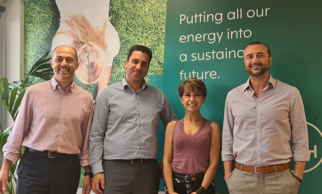 Four people stand smiling in front of a green wall with the text, Putting all our energy into a sustaining future. One woman and three men are dressed in business casual attire. A plant is visible on the left.