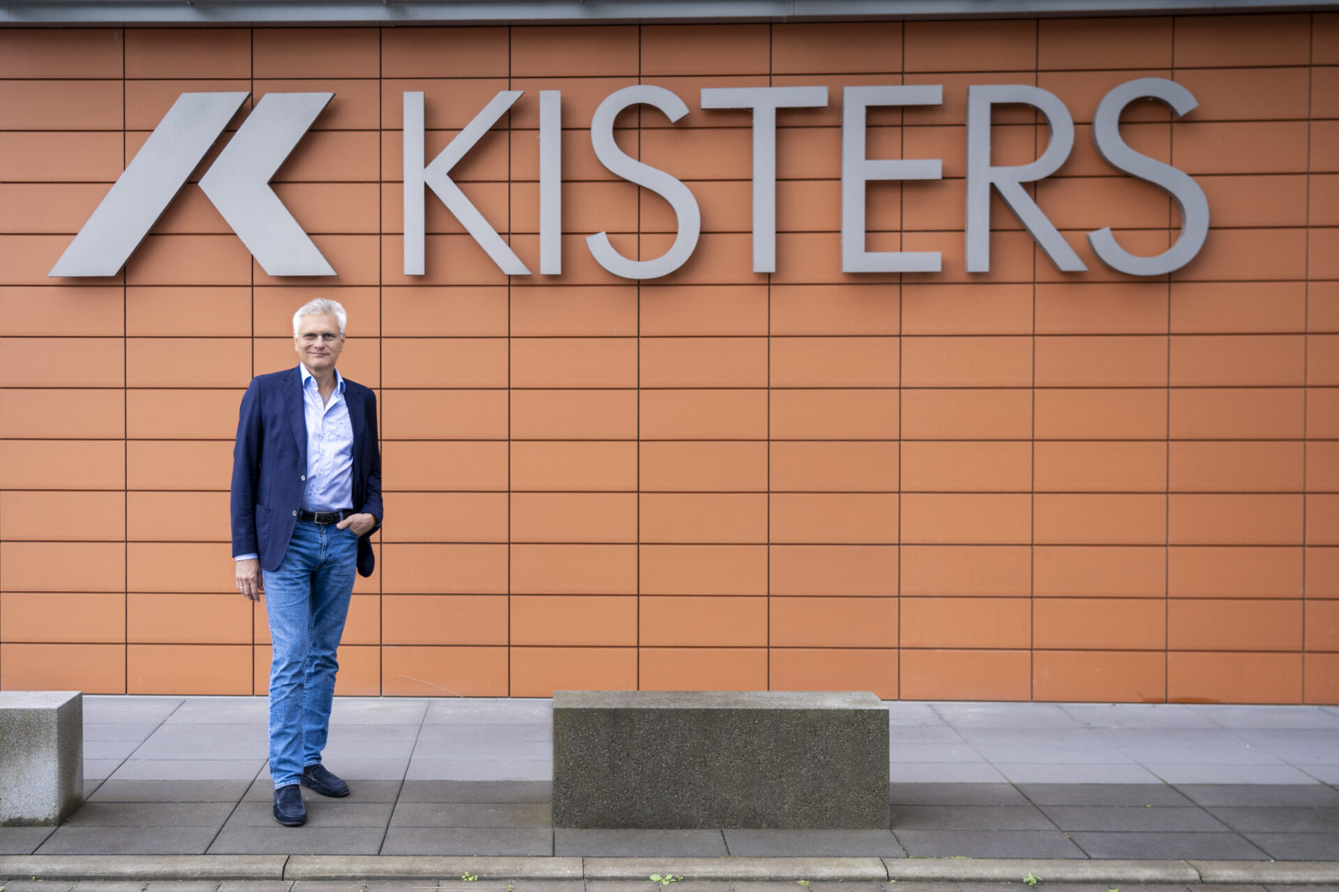 Ein Mann in einem blauen Blazer und Jeans steht vor einer orangefarbenen Kachelwand mit einem großen KISTERS-Schild und -Logo. Vor der Wand sind Betonbänke aufgestellt.