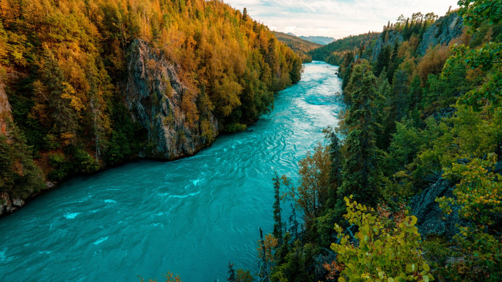Ein türkisfarbener Fluss fließt zwischen bewaldeten Hügeln mit herbstlich gefärbten Bäumen unter einem teilweise bewölkten Himmel und bildet eine malerische, natürliche Landschaft.