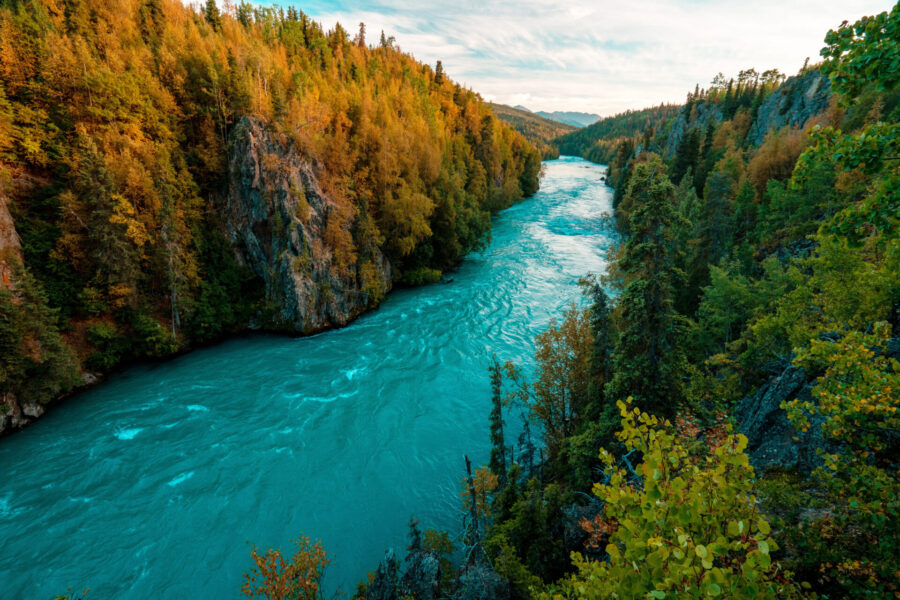Ein türkisfarbener Fluss fließt zwischen bewaldeten Hügeln mit herbstlich gefärbten Bäumen unter einem teilweise bewölkten Himmel und bildet eine malerische, natürliche Landschaft.