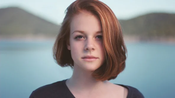 A young woman with shoulder-length red hair stands outdoors in front of a blurred lake and mountains, looking directly at the camera.
