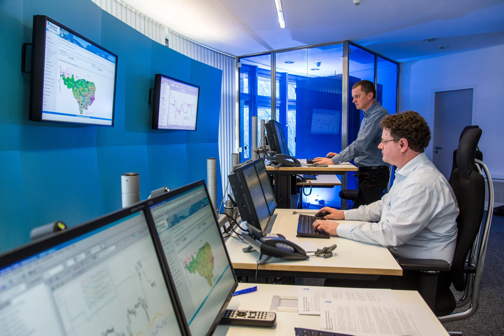 Two men work at computer desks in a modern office with large monitors showing maps and data charts. One is seated typing, the other stands at a high desk. The environment is bright, with blue accent walls and glass partitions.