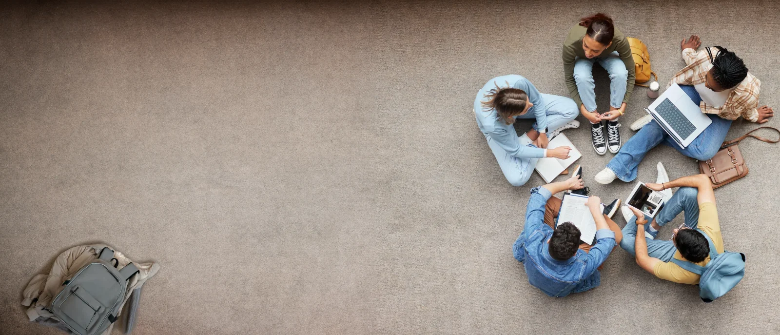 Fünf Schüler sitzen im Kreis auf einem Teppichboden und lernen gemeinsam mit Büchern, Heften, einem Laptop und einem Tablet. Ein Rucksack liegt allein auf dem Boden zur Linken. Die Gruppe scheint in eine Diskussion vertieft zu sein.