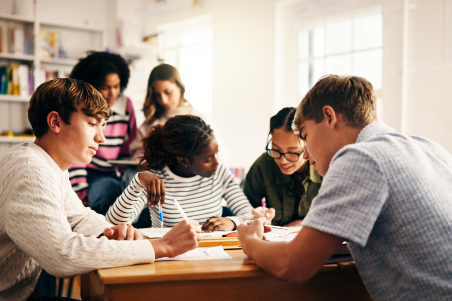 Eine Gruppe von Schülern sitzt in einem hellen Klassenzimmer um einen Tisch herum, arbeitet gemeinsam an Aufgaben und schreibt in Hefte. Einige andere sitzen im Hintergrund und sind ebenfalls auf ihre Arbeit konzentriert.